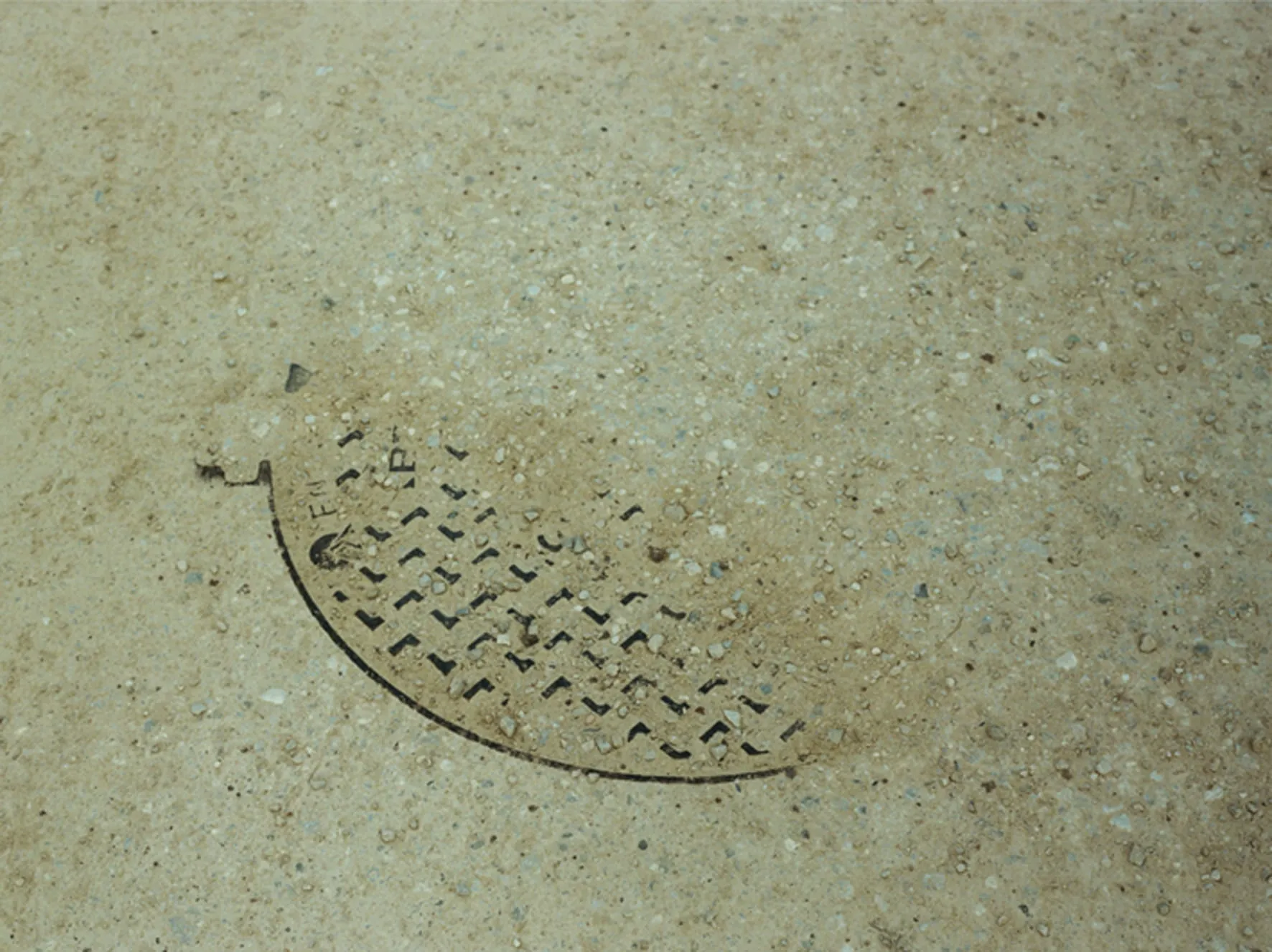 A semicircular metal drain grate half swallowed by a pale concrete floor, its perforated pattern still legible under a scatter of fine gravel.