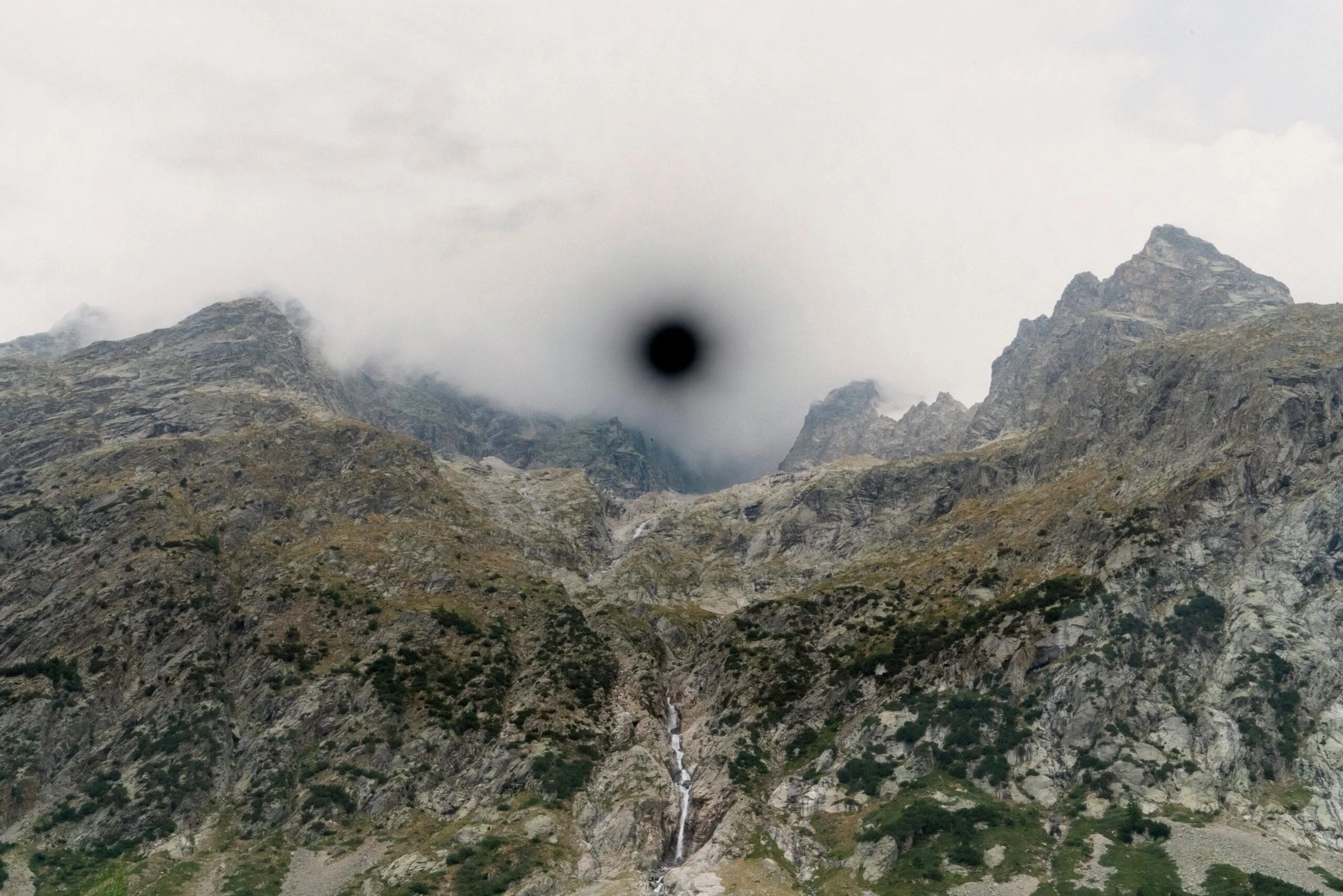 An alpine landscape of rocky peaks under low cloud, a thin waterfall running down the centre, with a black circular void burned into the sky and fading outwards like a dark halo.