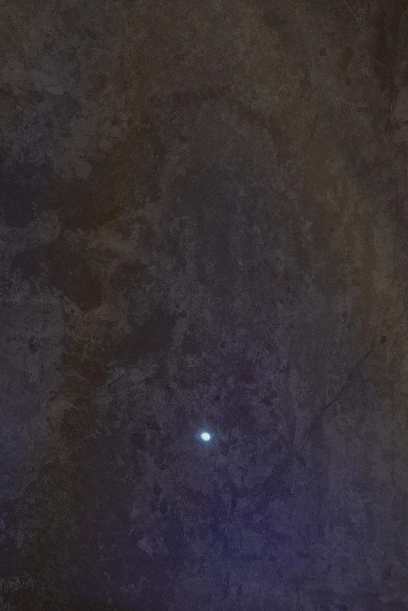 A mottled surface filling the frame, ochre and brown above shading to deep violet below, with a single small point of white light low and right of centre like a moon, and a faint diagonal crack running down the right edge.