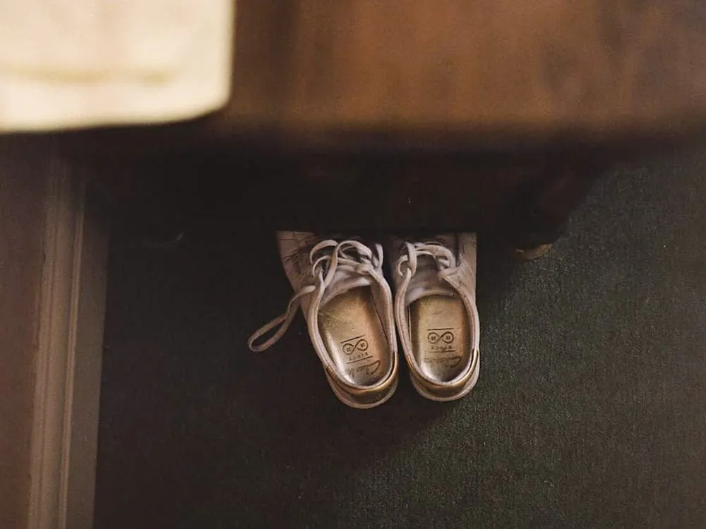 A pair of worn pale leather lace-up shoes left side by side on a dark green carpet, in the shadow beneath a wooden piece of furniture.