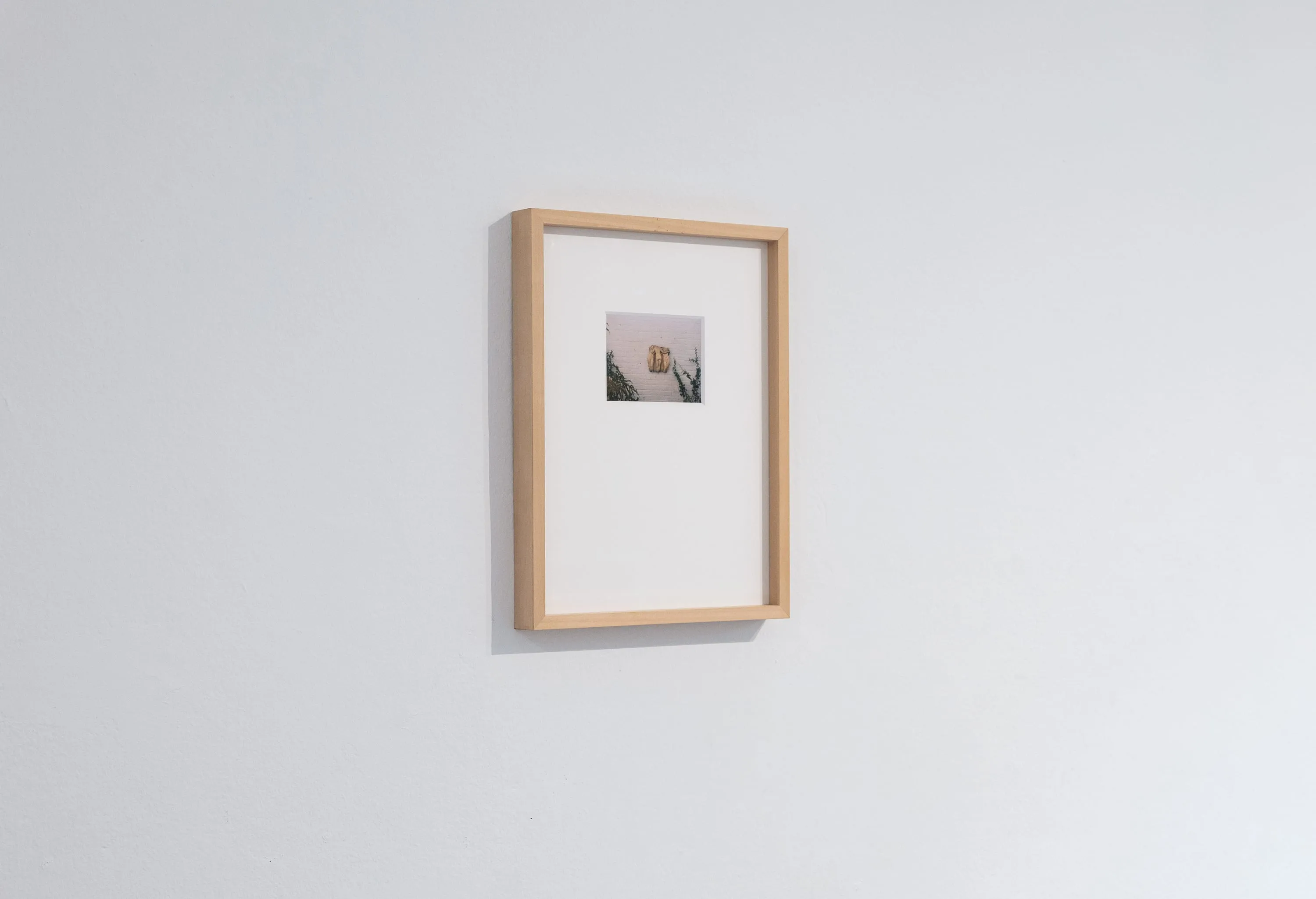 Installation view: the print is a small image set high in a wide white mount, in a pale wood frame on a white wall, seen at an angle.