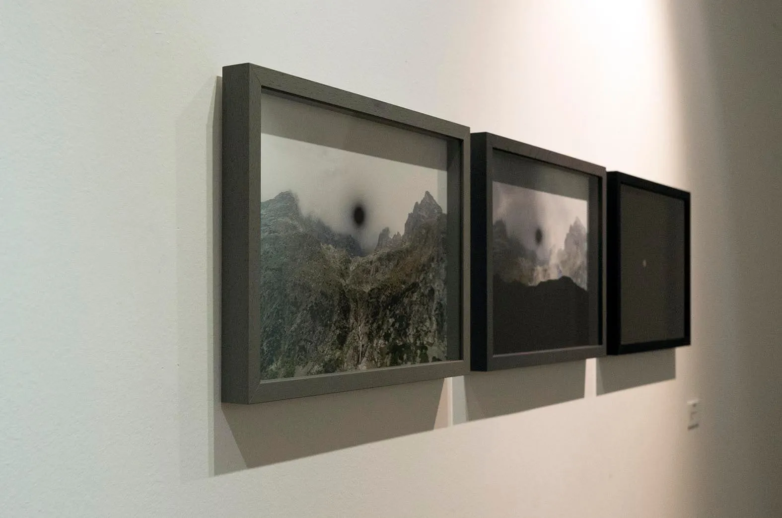Installation view: the three framed prints hung in a row on a white wall, each darker than the last, seen at an angle in raking light.